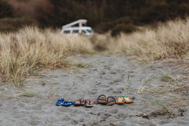 4 pairs of sandals sit on a beach with a white, pop-top camper van in the background.
