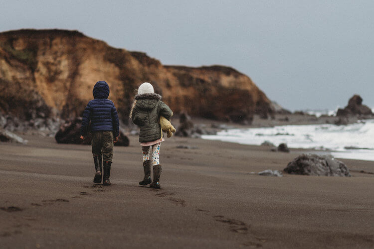 Two children walk alongside one another in winter coats on a beach,