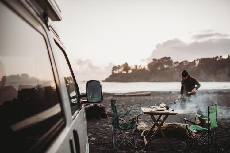Man cooks dinner on a camp table near a Volkswagen Eurovan Camper at the beach. 