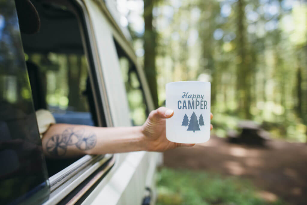 Woman holds Happy Camper cup out the window of a Volkswagen Vanagon Camper.