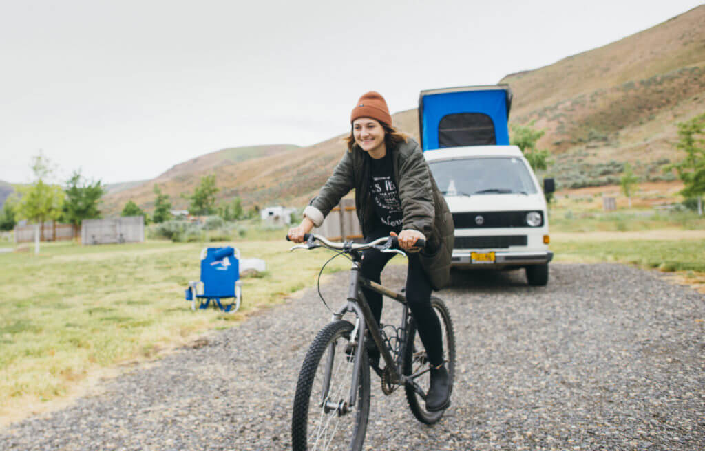 A woman with a brown hat and gray jacket rides a loaner bike in Cottonwood Canyon State Park while smiling with a white GoCamp camper van with a pop-top in the background