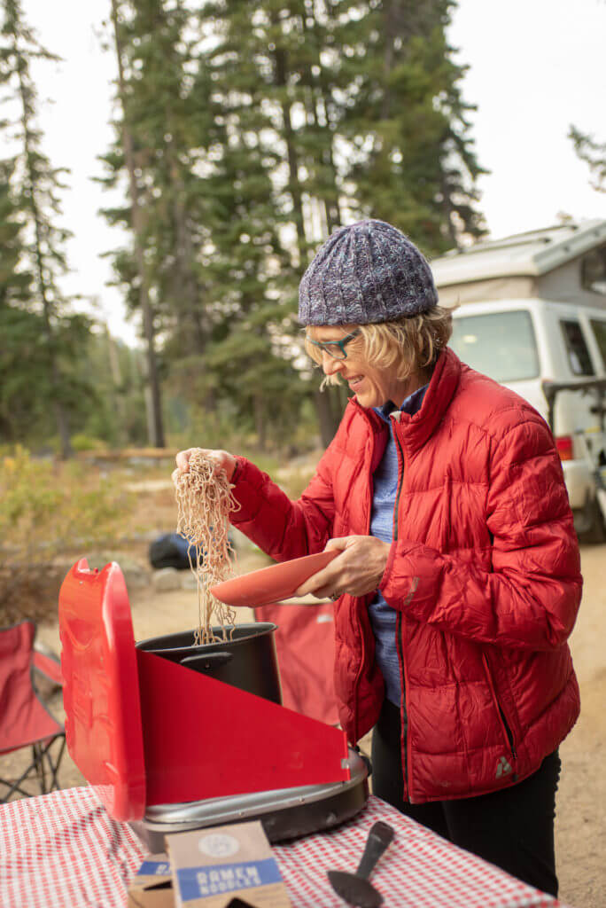 Woman in red coat puts ramen noodles in a bowl at a campground.